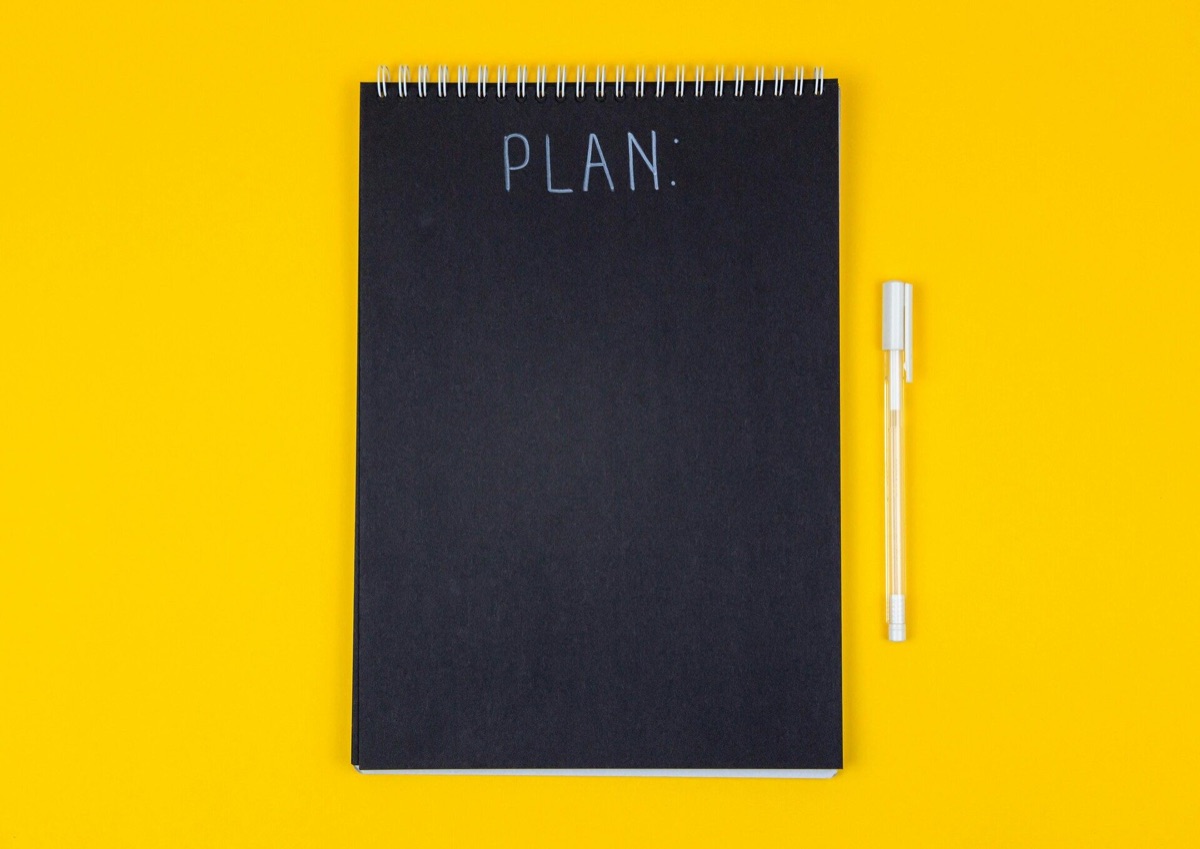 A black notepad labeled “PLAN:” with a pen on a yellow background